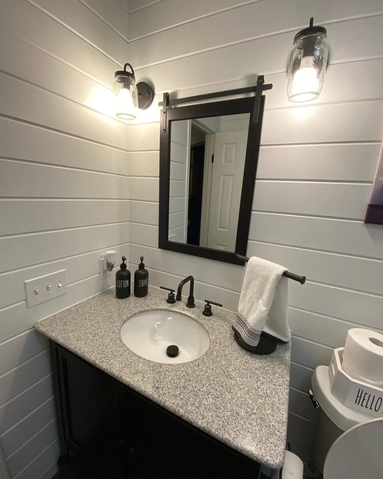 A half bath with white shiplap walls, a dark vanity with a granite top and black fixtures