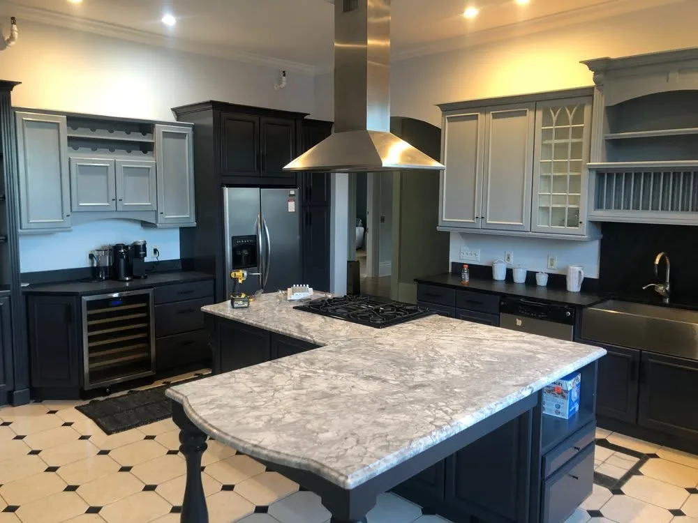 A large kitchen with dark cabinets and an island topped in white and gray stone under a steel range hood